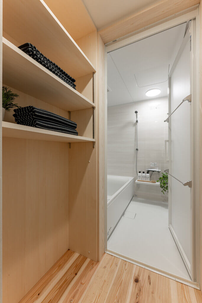 Wooden shelves with folded black towels beside a modern bathroom with a white bathtub and shower.