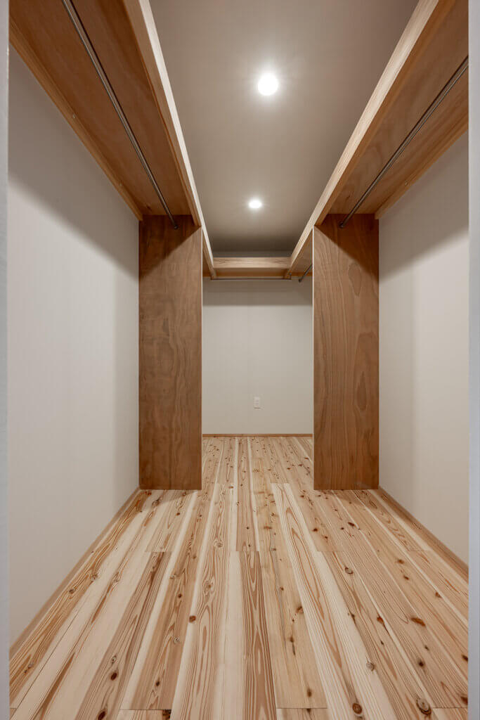 Narrow walk-in closet with light wood flooring, two vertical wooden pillars, and metal clothing rods along the walls under open shelving.