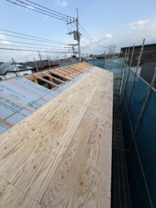 Wooden roof framing under construction with plywood sheathing on a scaffolded building, power lines in the background.