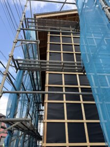Under-construction building with wooden framing and black weather barrier, surrounded by metal scaffolding and blue construction netting.